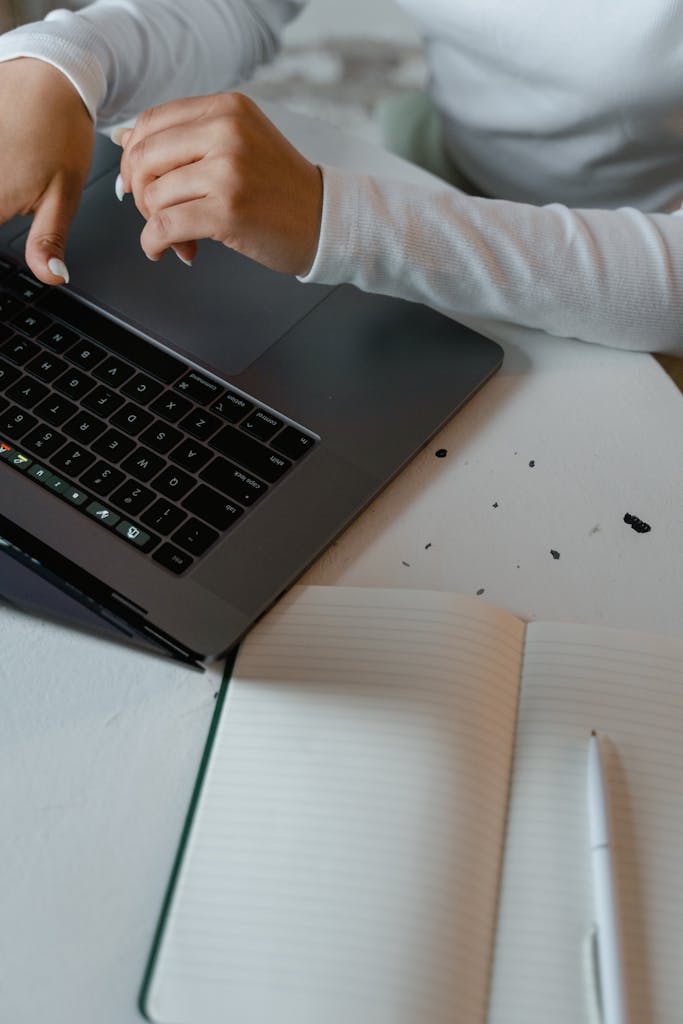Close-up of hands using a laptop with an open notebook nearby.